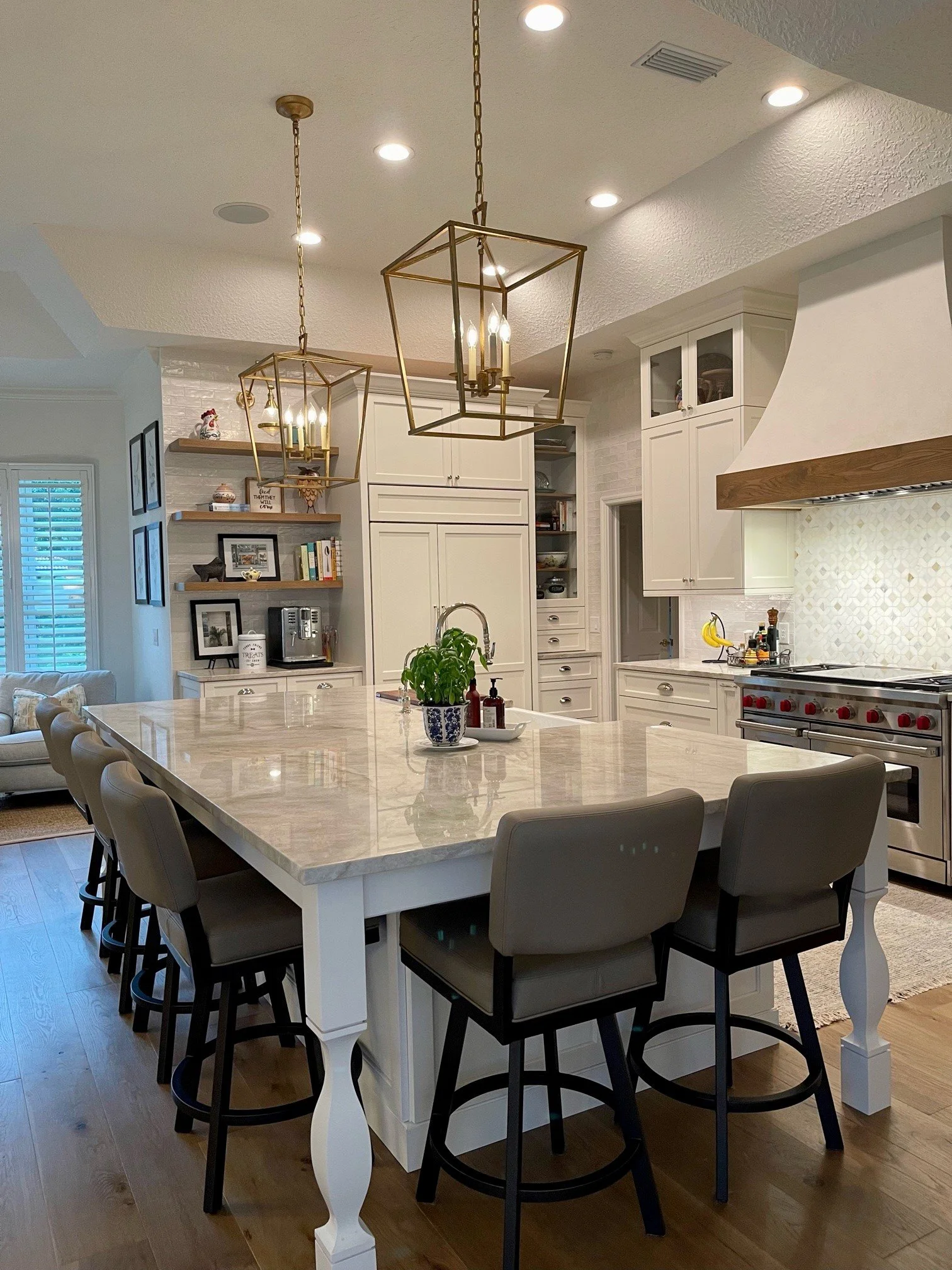A kitchen with white cabinets, a large marble island with beige chairs, pendant lights, a stainless steel stove, and decorative items on open shelves and the countertop.