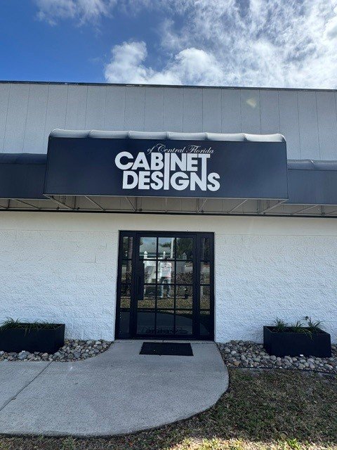 Storefront with a sign reading 'Cabinet Designs of Central Florida' above the entrance, which has a glass door and plants on either side.