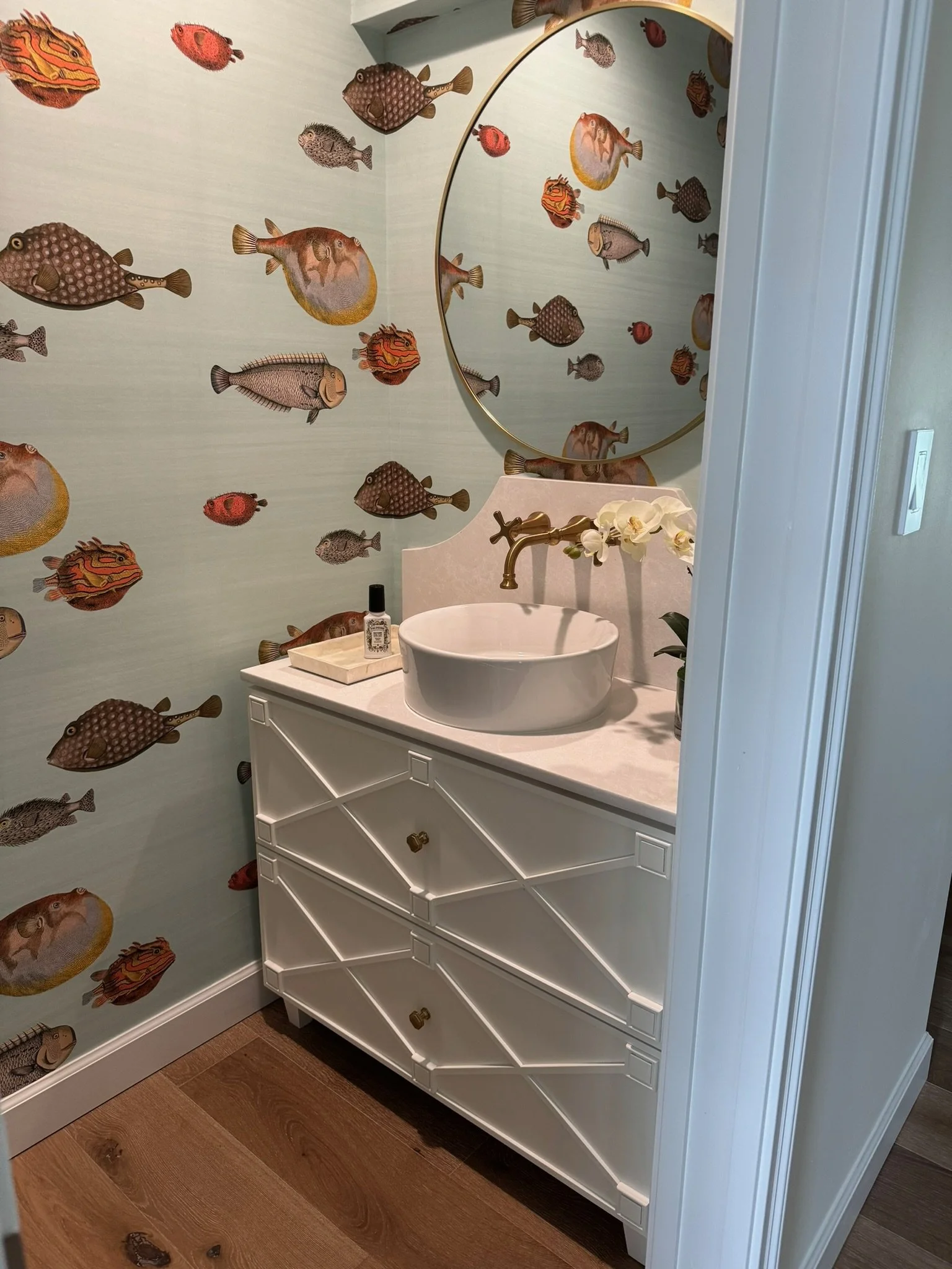 A bathroom vanity with a round vessel sink, gold faucet, white drawers, and a orchid plant on top. The wall is decorated with a wallpaper featuring various colorful fish.