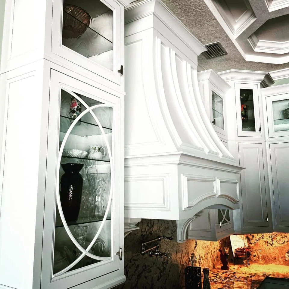Close-up of a white kitchen cabinetry with glass doors, intricate molding, and a granite backsplash illuminated with warm lighting.