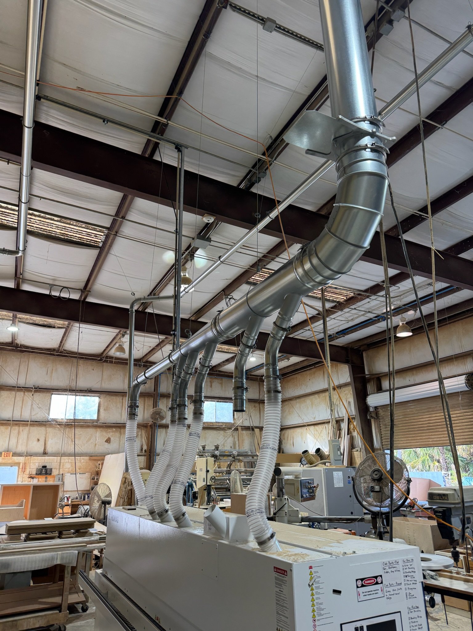 Industrial workshop with large metal ductwork and ventilation pipes hanging from the ceiling, various machinery, workbenches, and shelves, with windows letting in natural light.