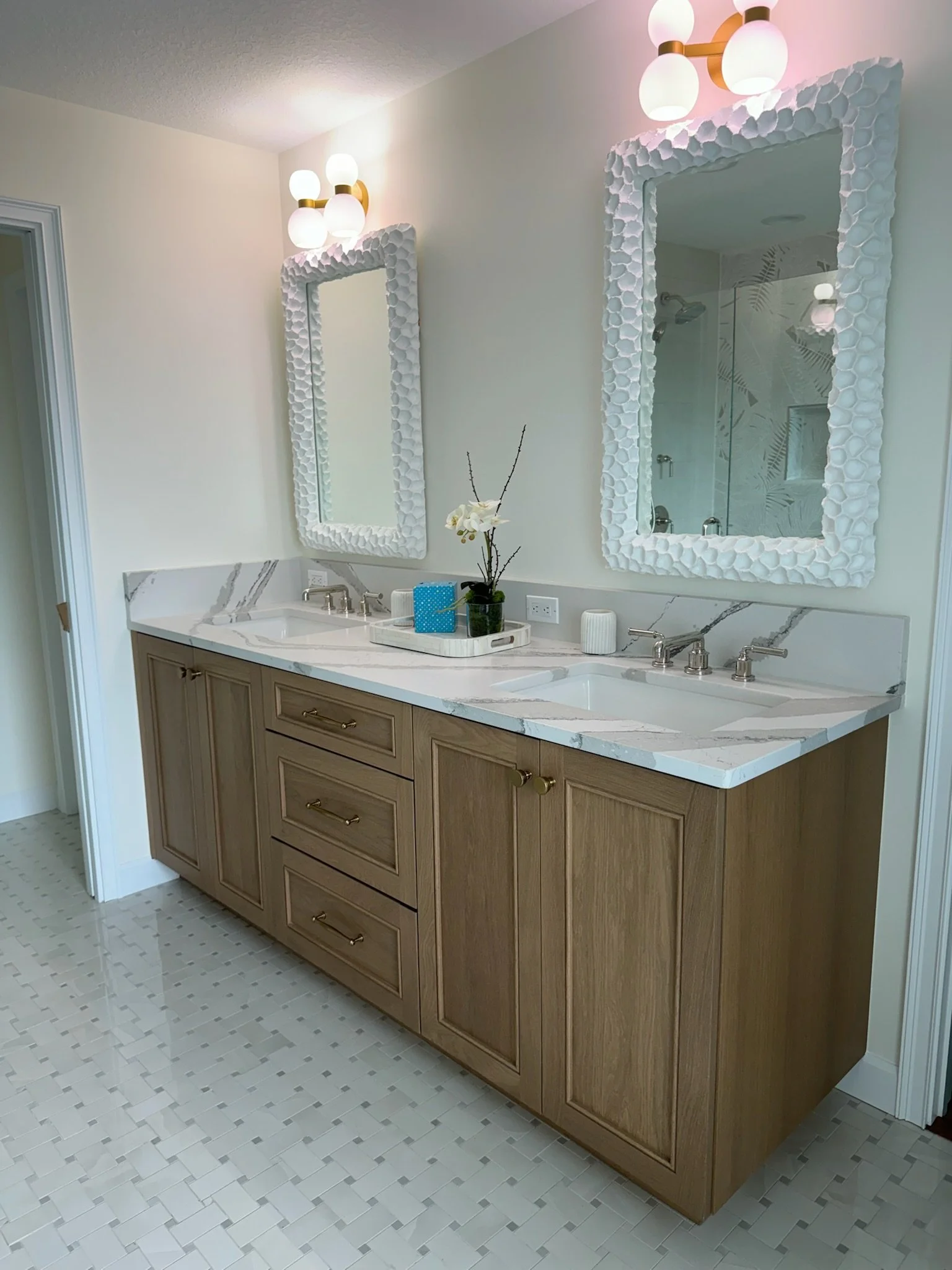 Bathroom with two mirrors featuring textured white frames, a marble countertop with a double sink, a light-colored wooden vanity, and a floor with small square tiles. There is a decorative tray with a plant and other items, and modern light fixtures 