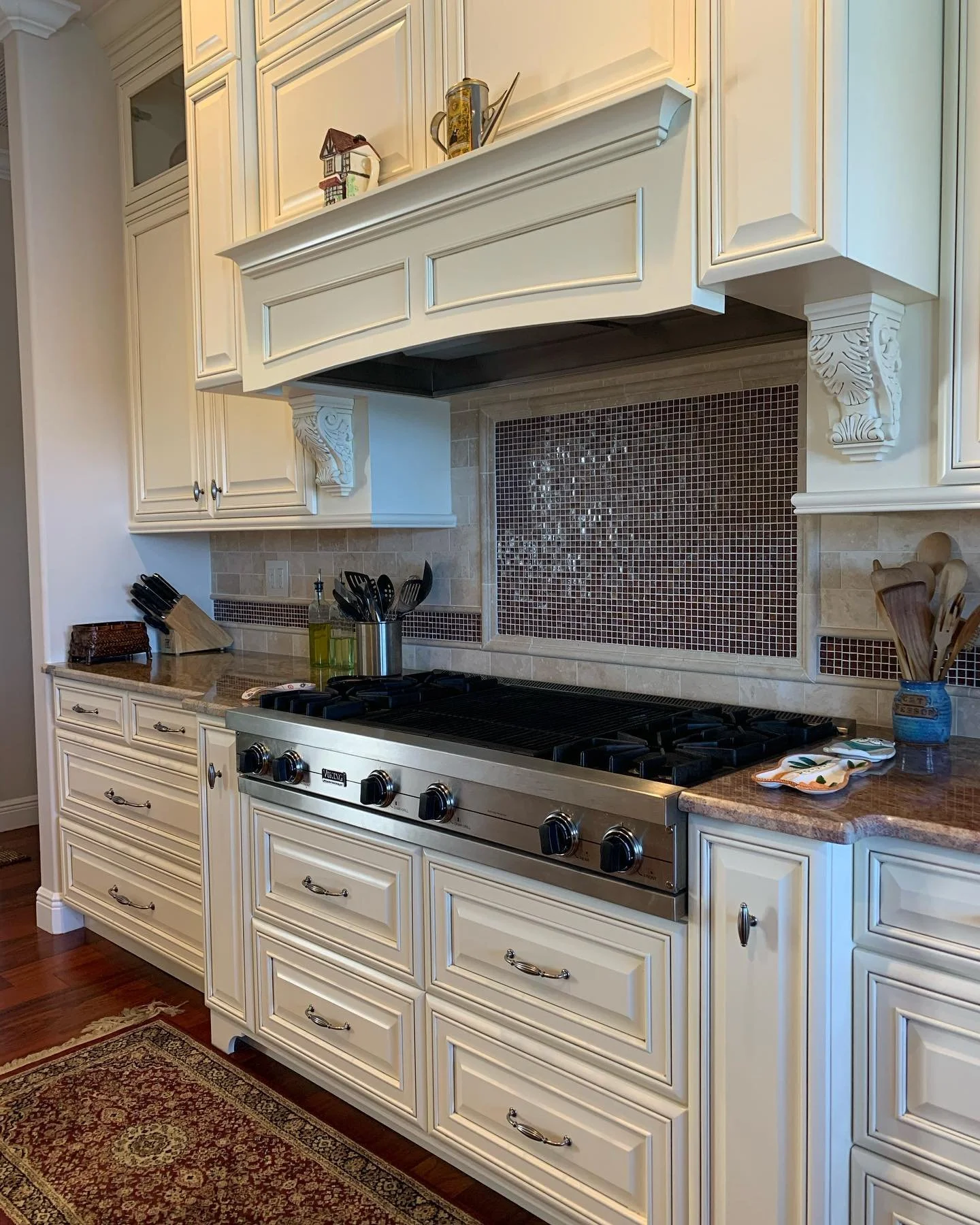 Cream-colored kitchen cabinets above a granite countertop with a stove. Decorative corbels support a range hood with a mosaic tile backsplash behind the stove. Kitchen utensils and a small dish are on the countertop, and a patterned area rug is on th
