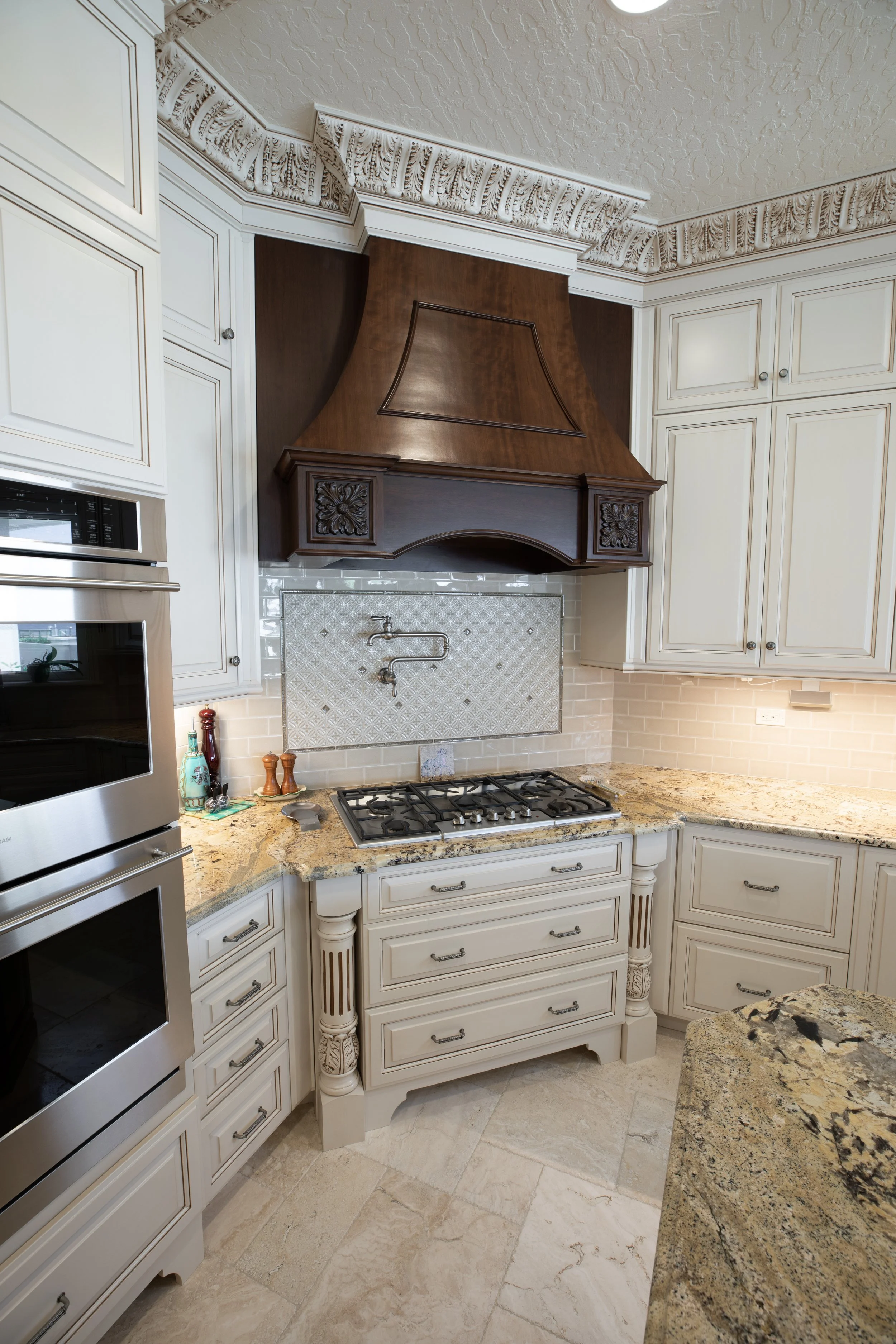 A kitchen with beige tile flooring, cream-colored cabinets with decorative moldings, granite countertops, a stainless steel oven, and a wooden range hood with ornate carvings.