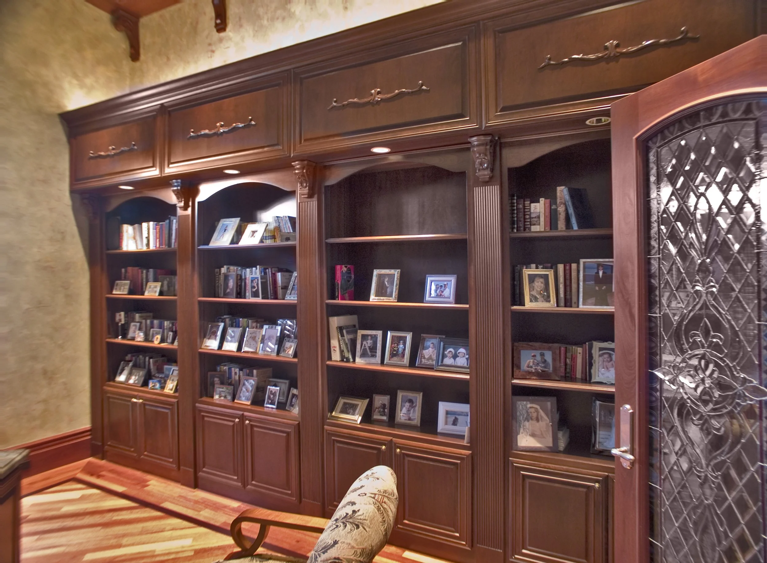 Wooden bookshelf filled with framed photographs and books, situated against a beige wall with decorative molding and patterned tiles on the right side.