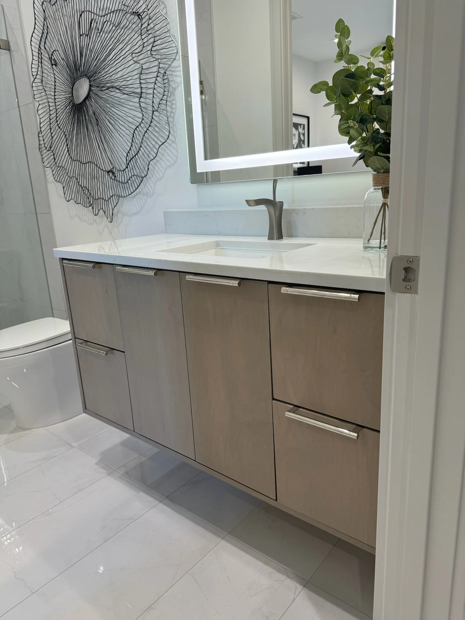 Modern bathroom with a white marble countertop vanity, a rectangular mirror with backlighting, a potted plant in a glass jar, a decorative wire wall art, and a toilet partially visible on the left.