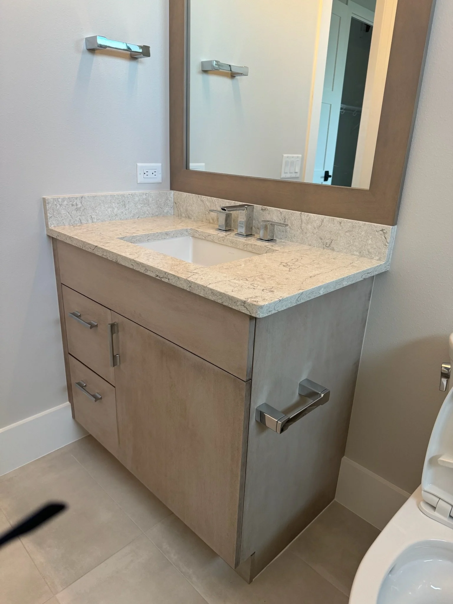 Bathroom vanity with a large mirror, beige marble countertop, sink, and chrome faucet. There are cabinets below, and a wall outlet beside the mirror. Part of a toilet is visible to the right.