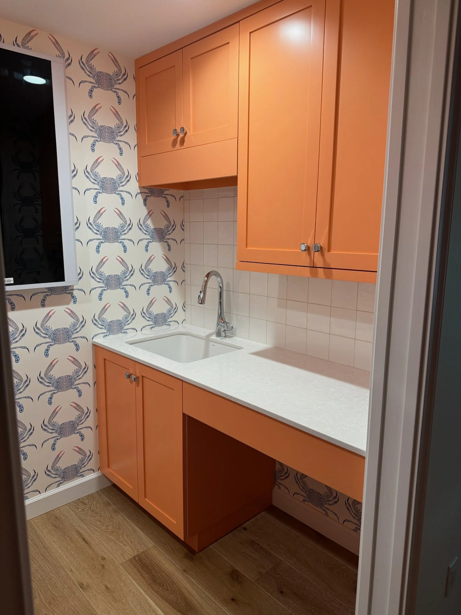 A kitchen with orange cabinets, a white countertop, a white tiled backsplash, a sink with a silver faucet, and wallpaper with blue and red crab designs.