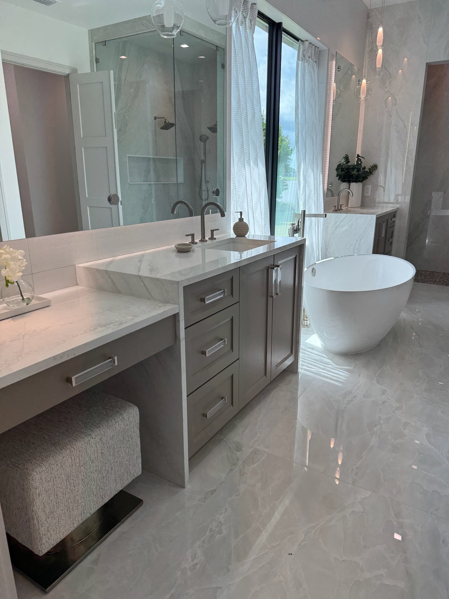 Modern bathroom with a free-standing bathtub, a vanity with a sink and decorative items, a shower area with glass enclosure, large floor tiles, and a window with curtains.