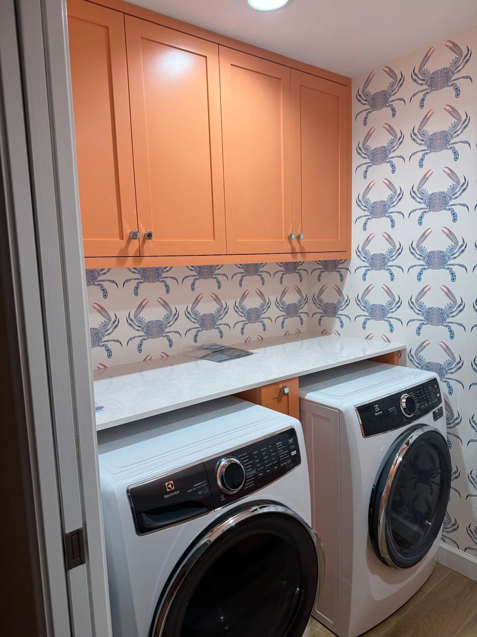 A laundry room with a white washing machine and a white dryer side by side below a white countertop. Above the appliances, there is a wooden cabinet with three doors. The wall behind features blue and gray crab-patterned wallpaper.