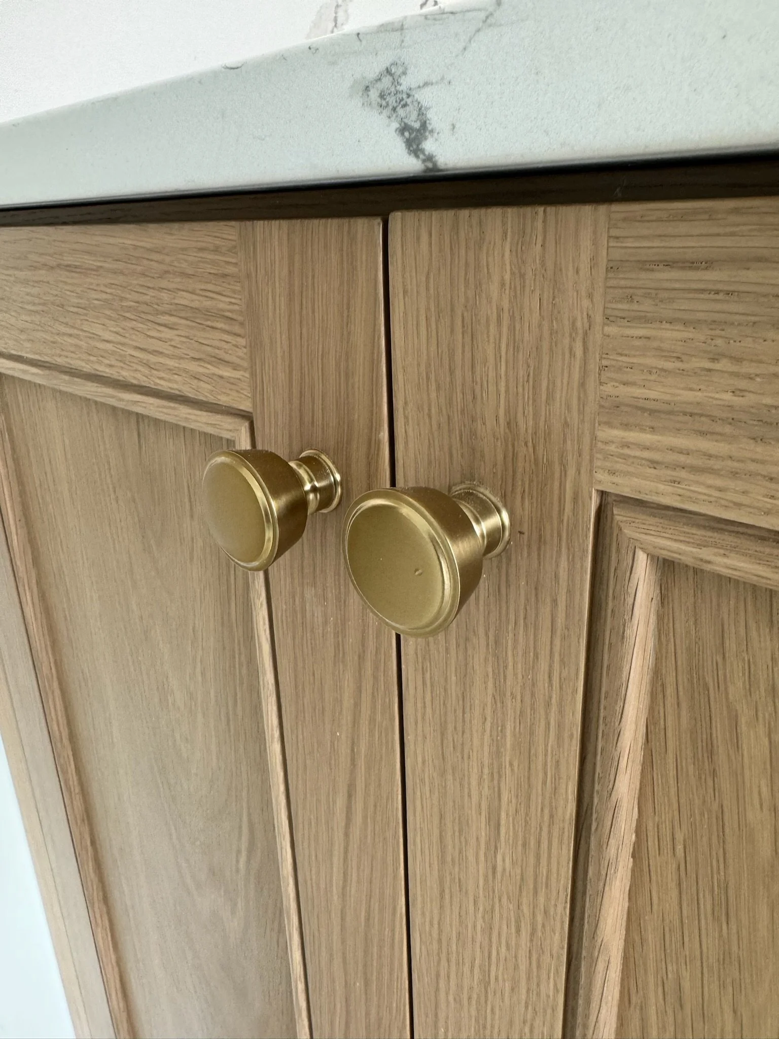 Close-up of a wooden door with two round, gold-colored door handles and a white wall above it, which has a dark smudge or stain.