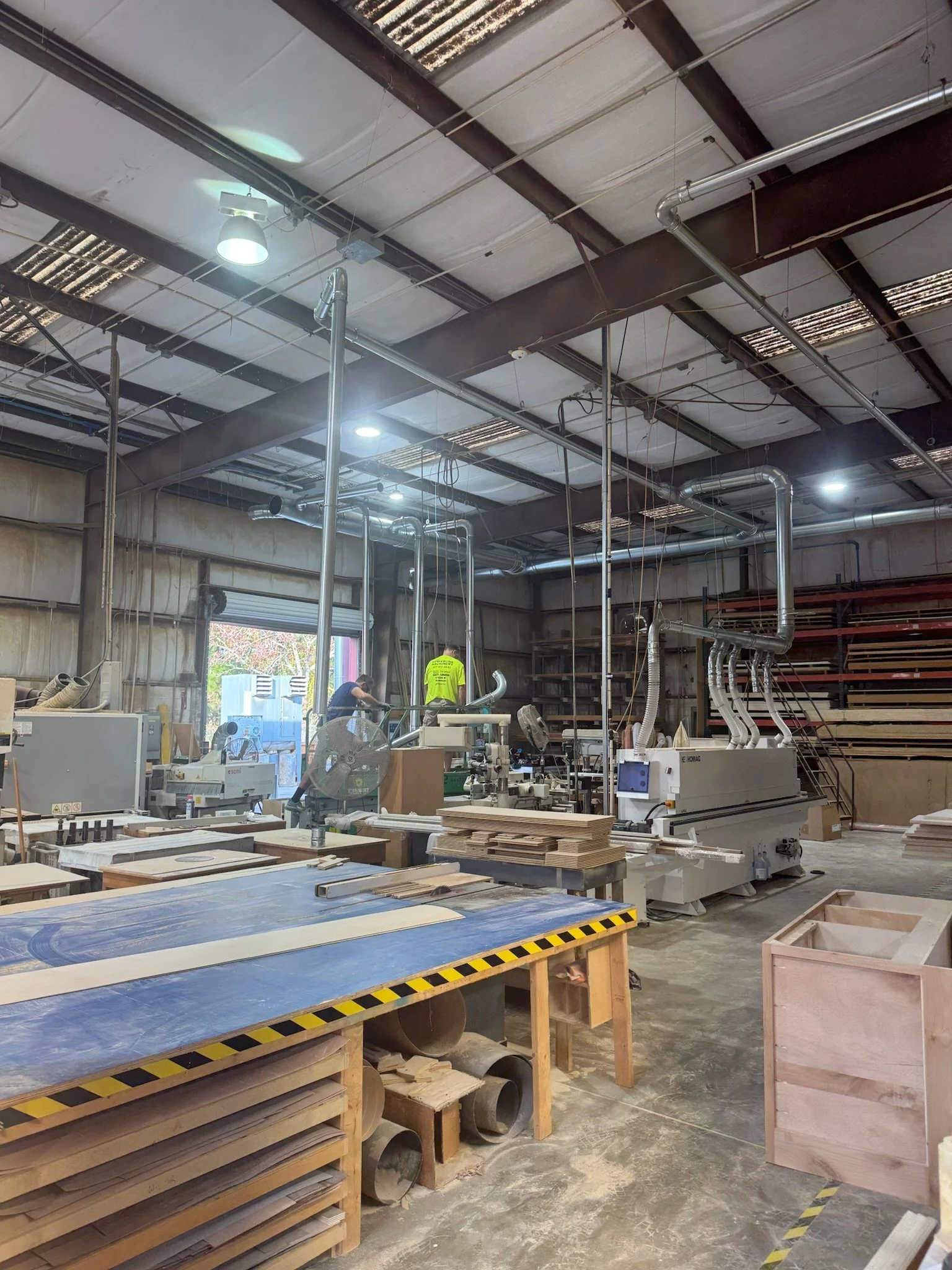 Interior of a woodworking or carpentry workshop with wooden furniture, tools, and pipes. Two workers are seen working, and sunlight enters through an open door.