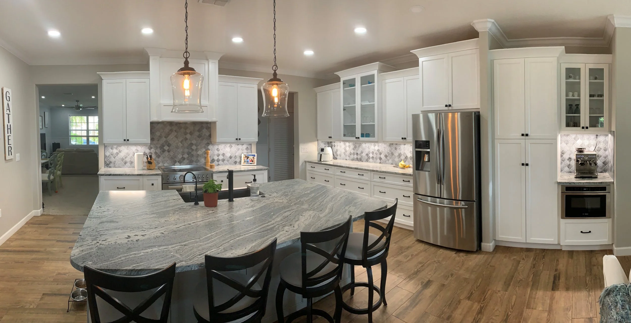 Modern kitchen with white cabinets, granite countertops, stainless steel refrigerator, and a large island with a granite top. Two hanging pendant lights over the island, and a view into a dining area and living room.