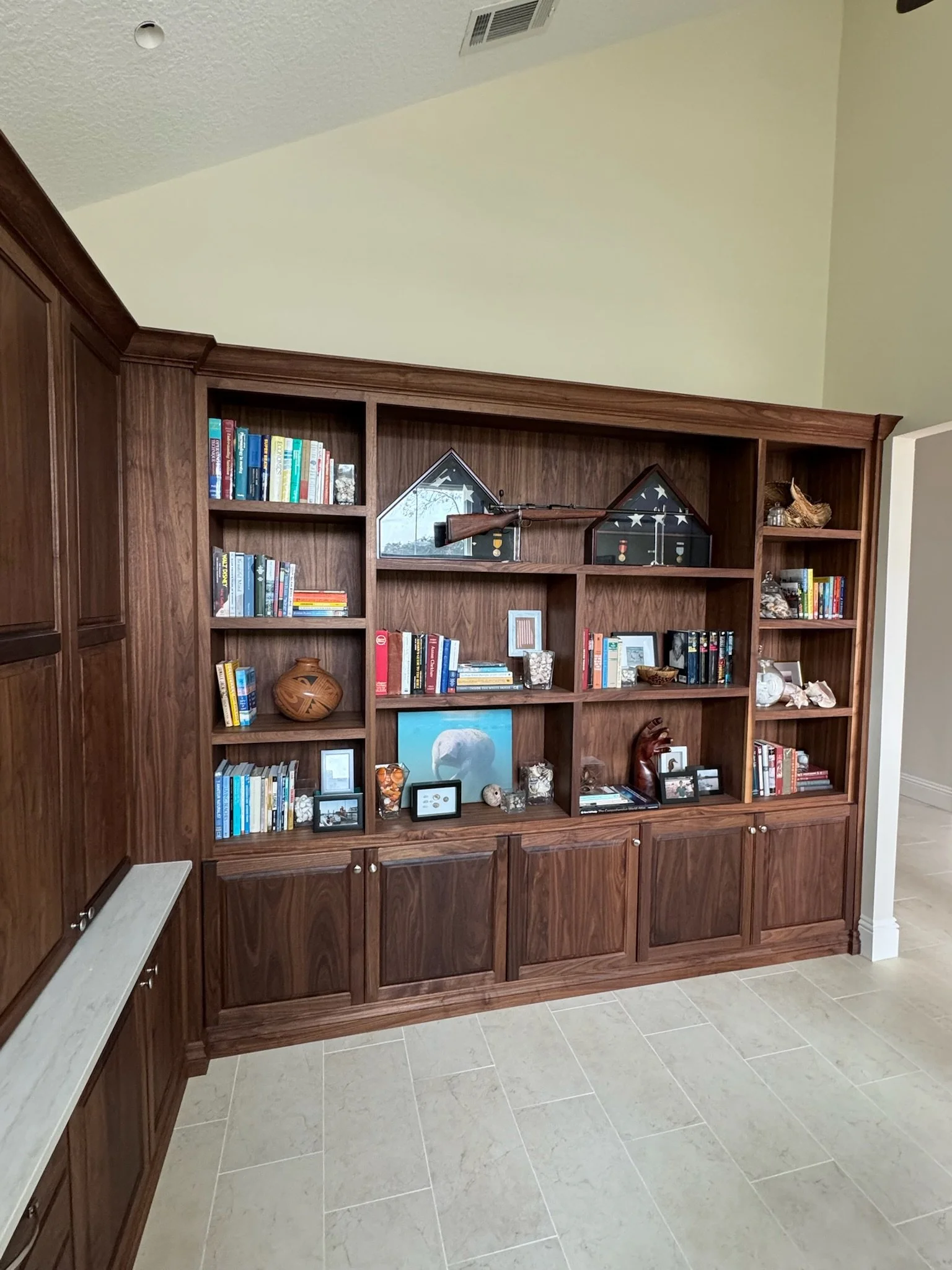 A large wooden bookshelf filled with books, decorative items, framed photographs, and artifacts, situated in a room with cream walls and light-colored tile flooring.