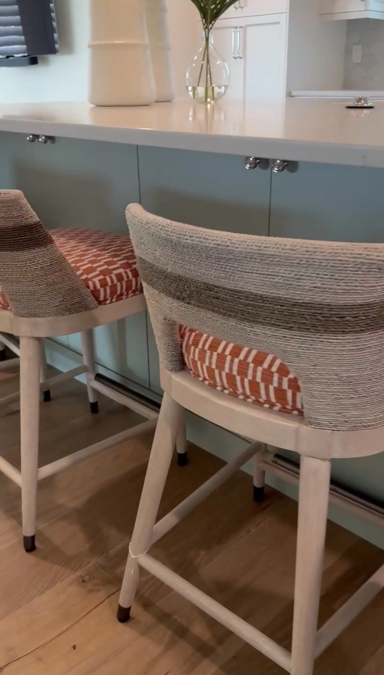 Two wooden chairs with woven fabric seats and backs, positioned at a kitchen counter. The chairs have plaid-patterned cushions. On the counter, there are decorative vases and a glass vase with green leaves.
