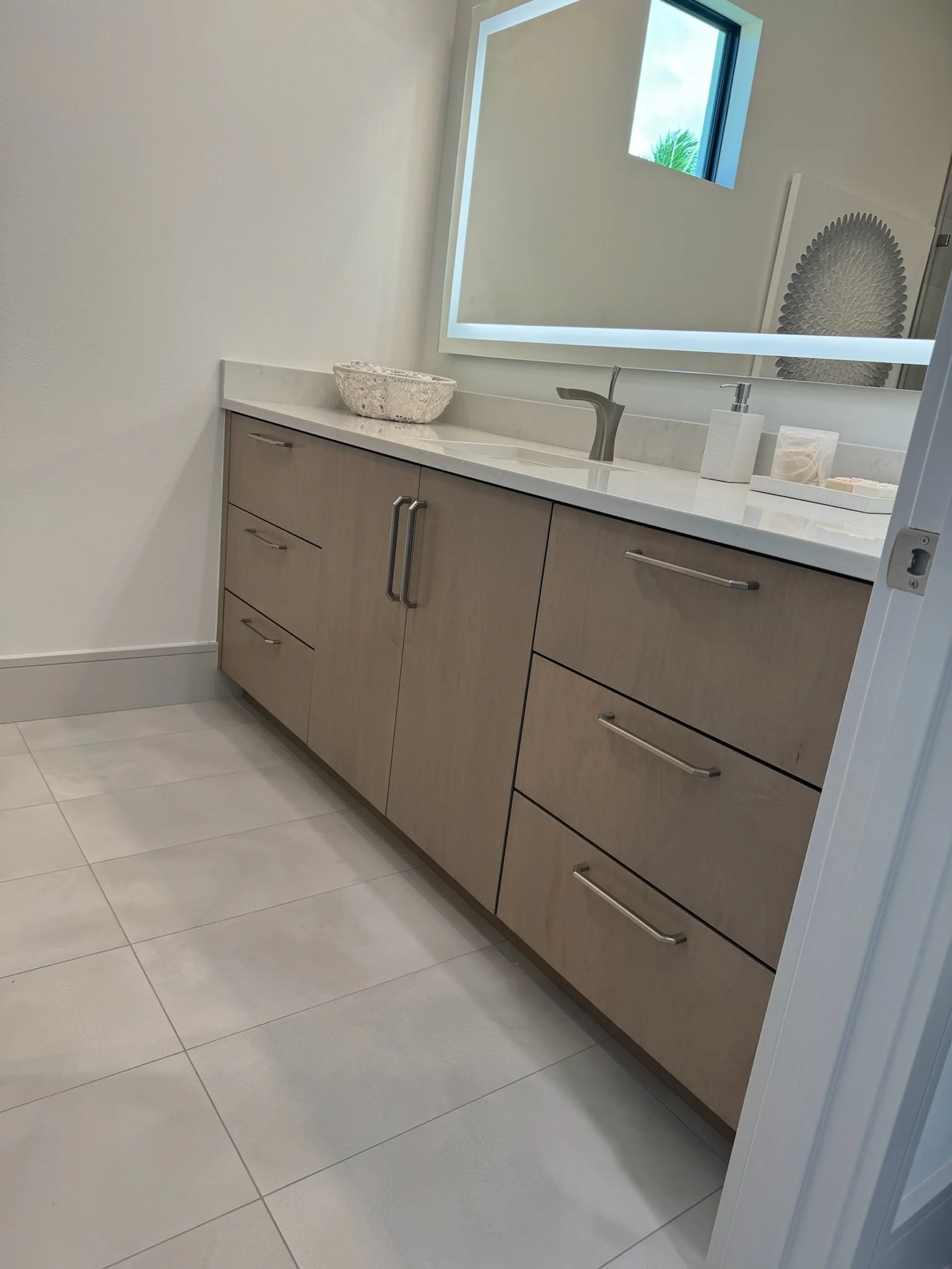 A bathroom vanity with a light wood cabinet, white countertop, and silver handles. There is a woven basket, a soap dispenser, and some small containers on the counter. A large illuminated mirror hangs above the sink, and a window with blue sky and gr