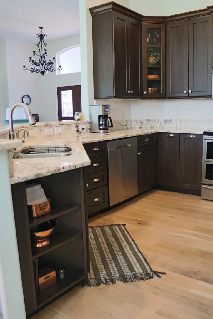 A modern kitchen with dark wooden cabinets, a white and gray marble countertop, stainless steel appliances, a black chandelier, a woven rug on light wooden flooring, and a window with a decorative glass panel.