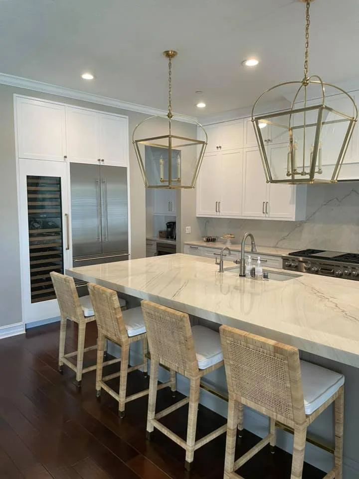 Modern kitchen with white cabinets, marble backsplash, large marble island, stainless steel appliances, and four wicker bar stools.