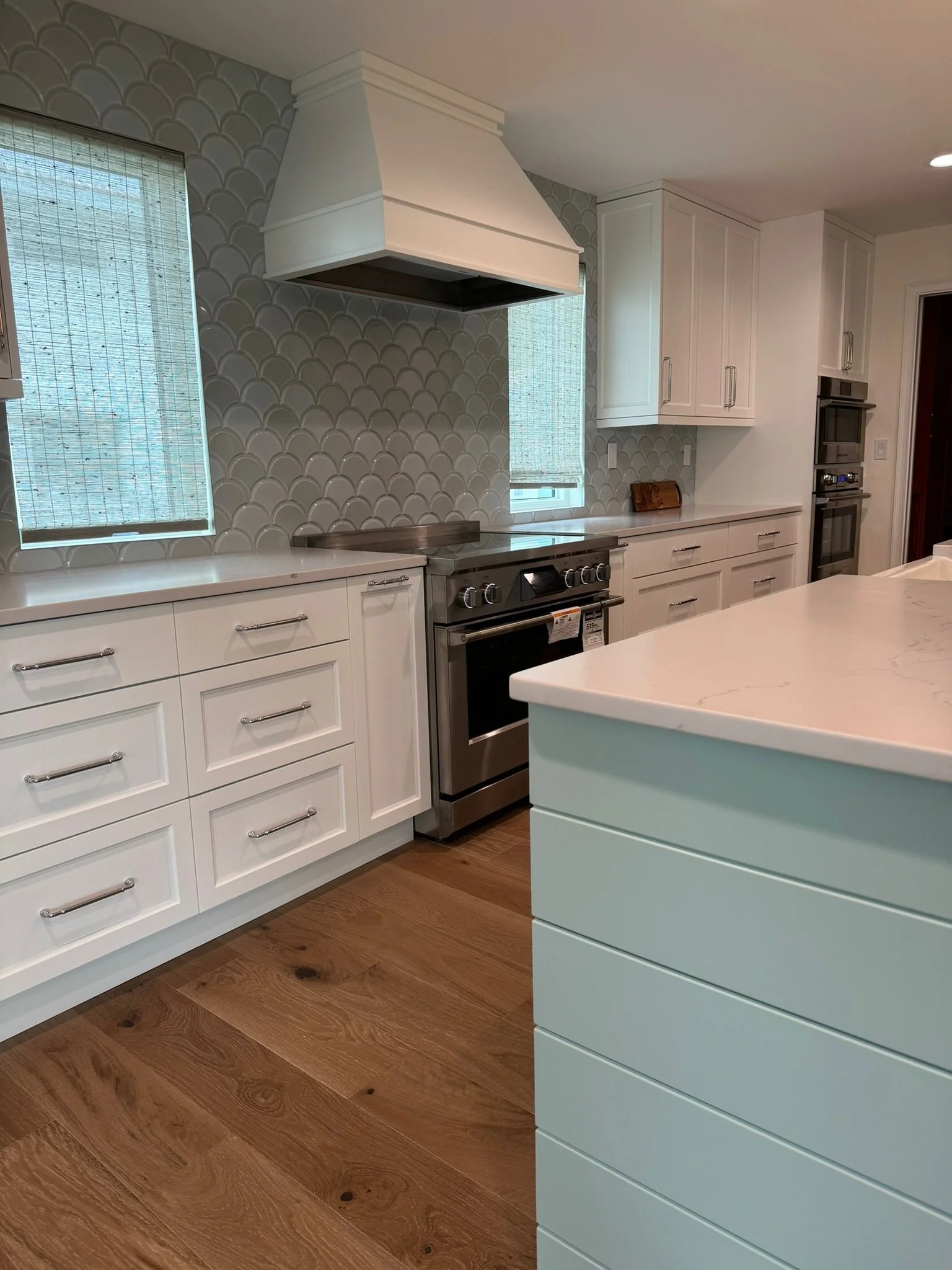 Modern kitchen featuring white cabinetry, a stainless steel oven, a white range hood, patterned glass backsplash, and wooden flooring.