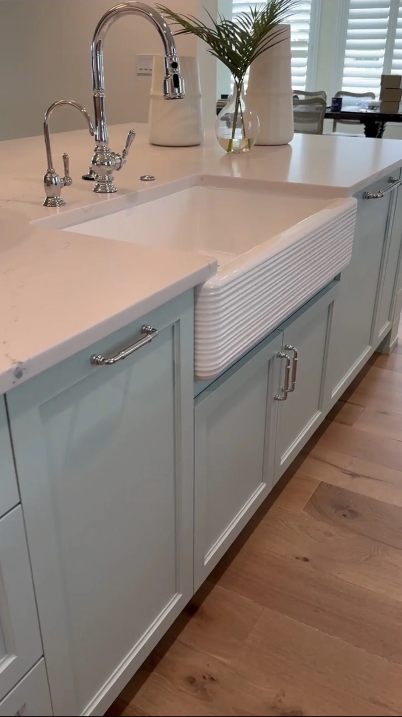 A kitchen sink with a white basin and a vintage-style faucet, set in a light blue cabinet with silver handles. On the counter, there are white vases with green plants and a glass vase with water and a green plant stem. In the background, there are wh