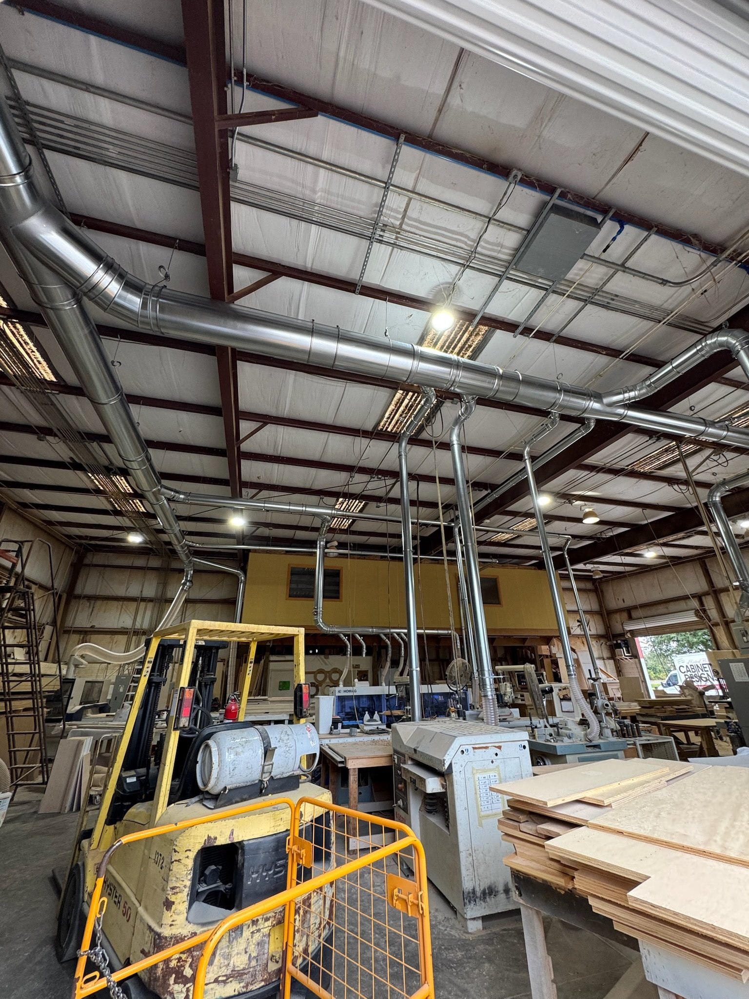 Indoor woodworking shop with various tools, machine, stacks of lumber, and ventilation pipes on the ceiling.