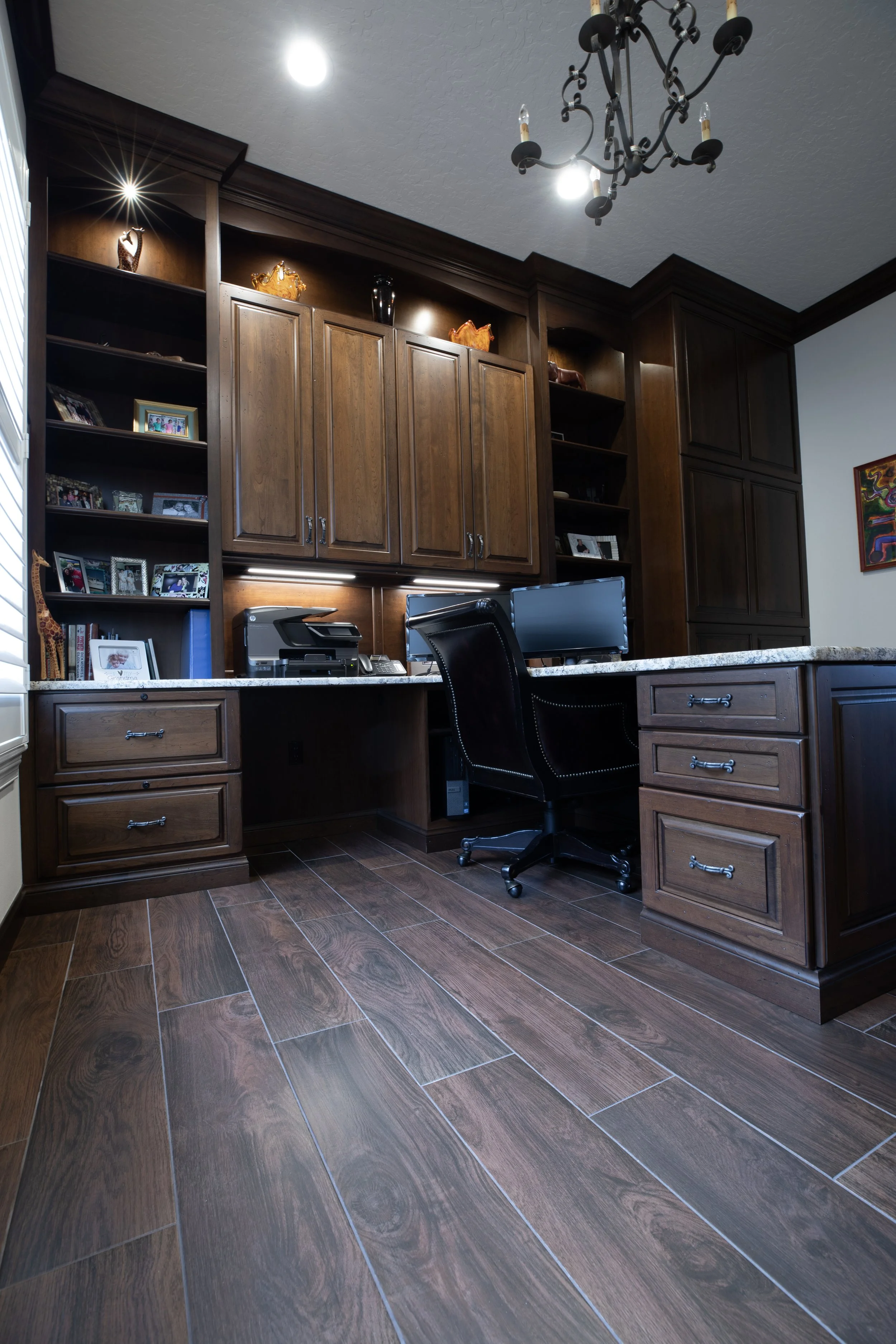 Home office workspace with wooden cabinetry, a countertop with a computer monitor, printer, and office supplies. Framed photos and decorative items are displayed on open shelves. The flooring is wood, and there is a black office chair. The ceiling ha