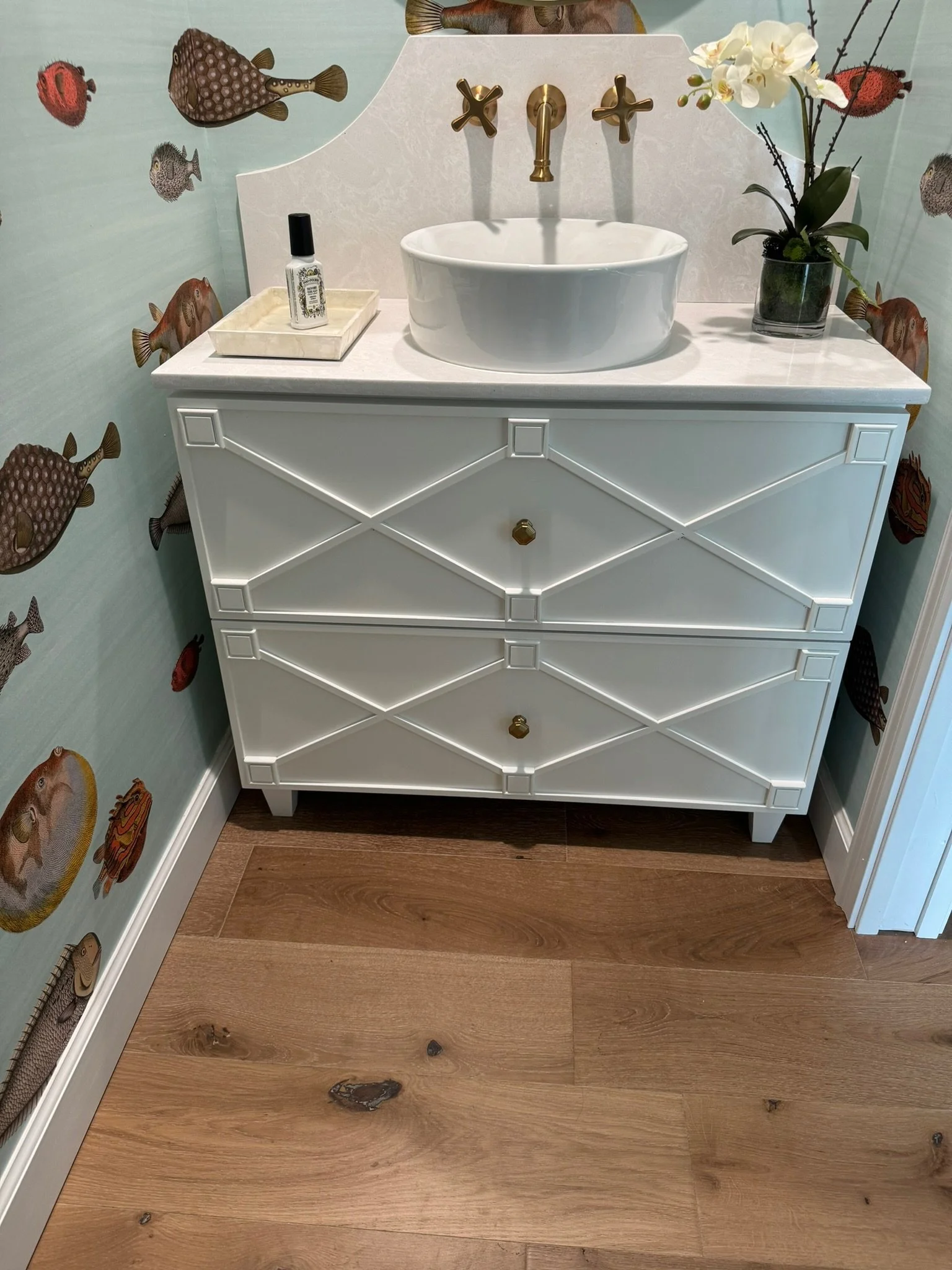 A white dresser with three drawers, featuring a gold knob on each drawer, placed in a room with light wooden floor. On top of the dresser is a white ceramic sink, a tray with nail polish, and a black vase with white orchids. The background wall has a