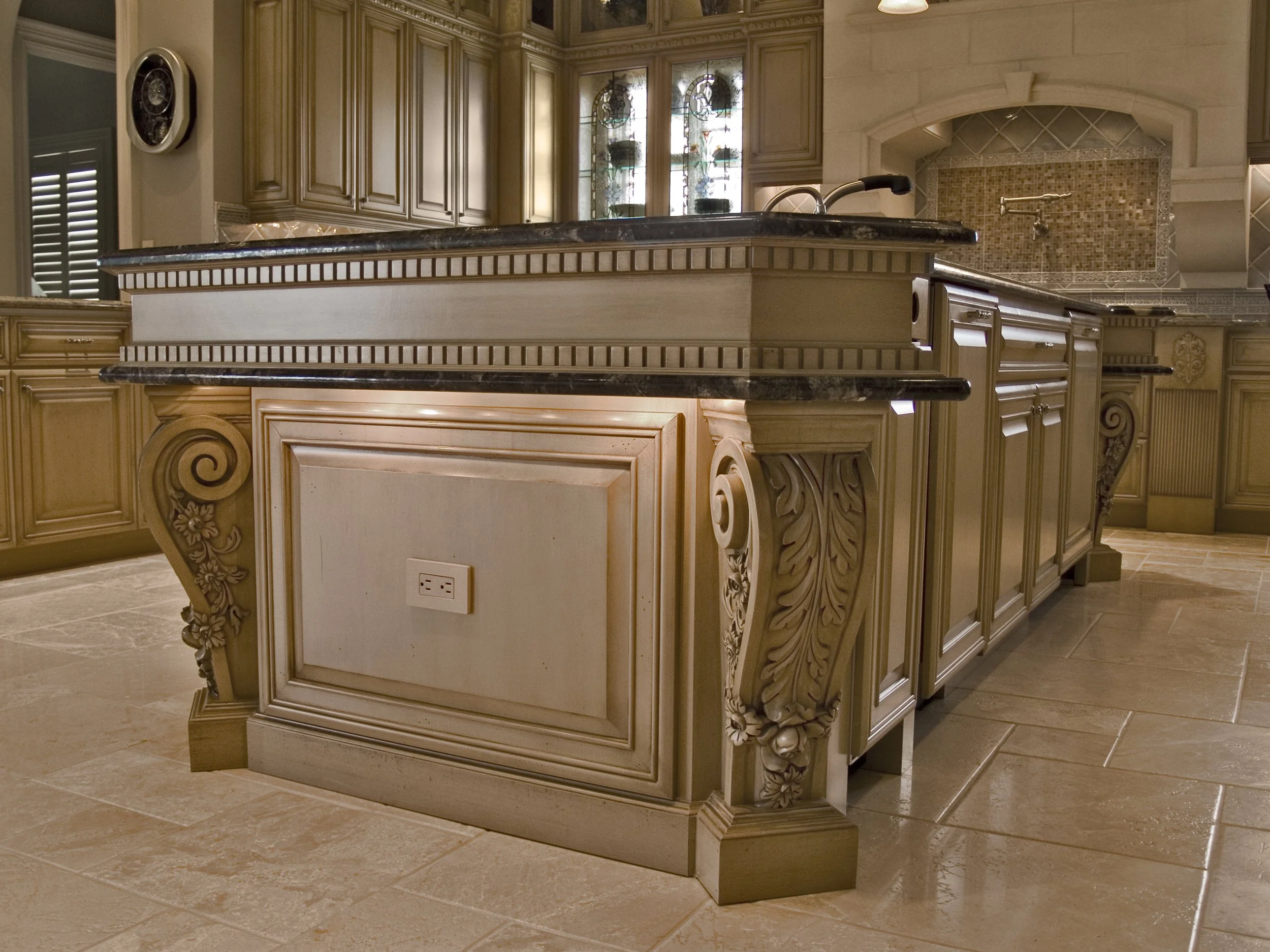 A kitchen with ornate wooden cabinetry, a marble countertop, a tiled backsplash under an arch, and a stained glass cabinet. Beige tiles cover the floor.