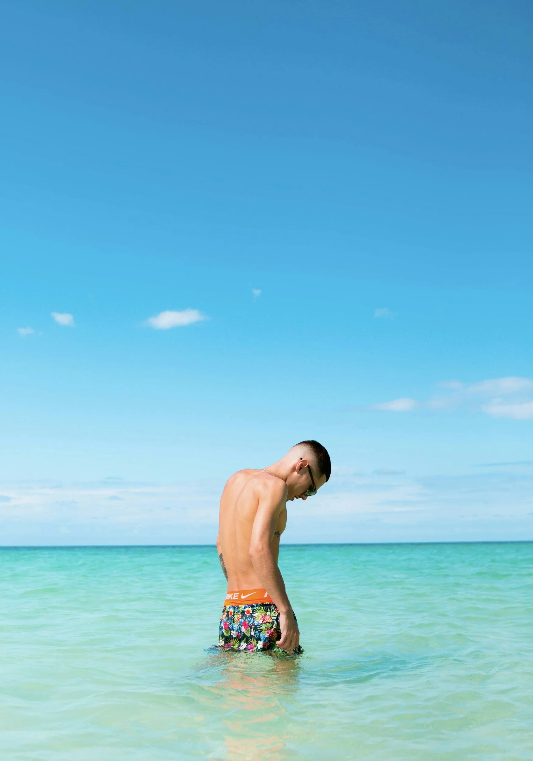 A shirtless man with sunglasses standing in shallow clear water at the beach, wearing colorful shorts with a floral pattern, with a blue sky and distant horizon in the background.