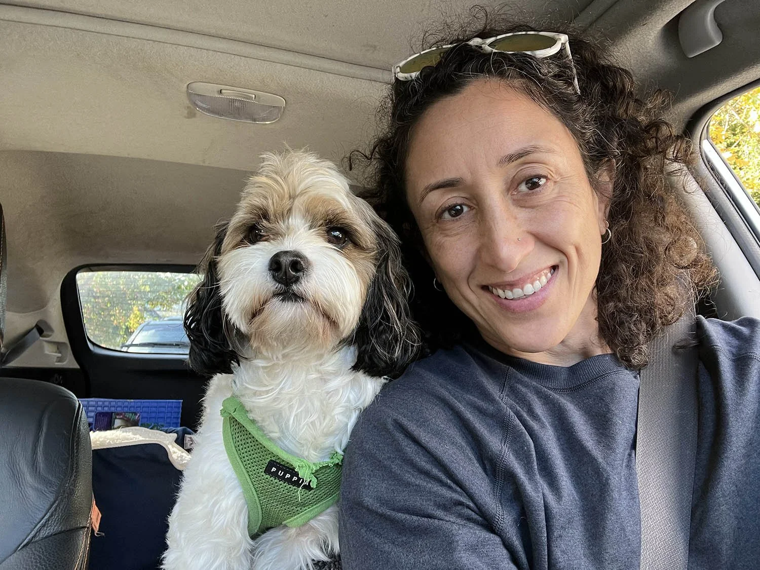 Woman and dog taking a selfie inside a vehicle. The woman is smiling, wearing a dark shirt, and sunglasses on her head. The dog, a fluffy white and brown breed with a green harness, sits next to her. Owner of Amala Studio in Lynnwood WA.