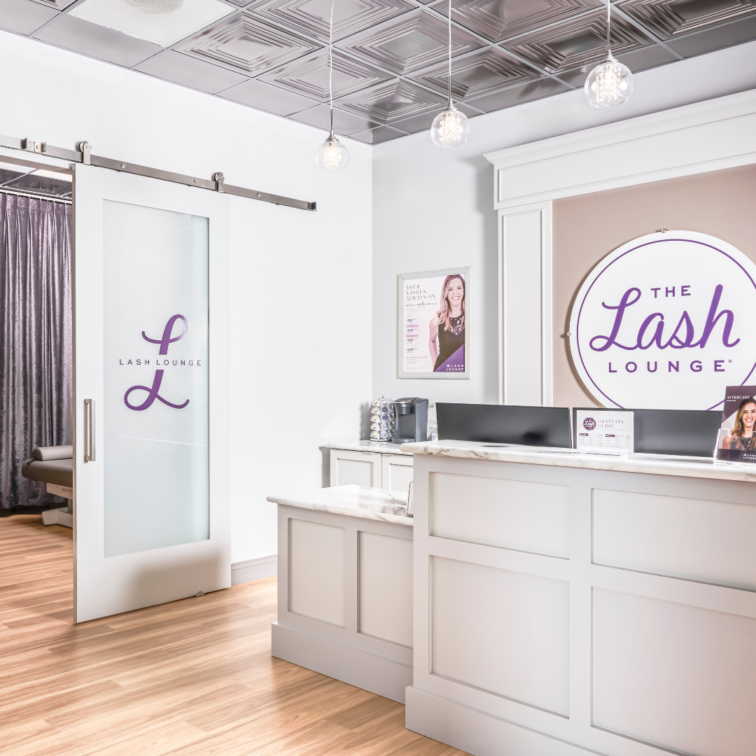 The reception area of The Lash Lounge salon featuring a white counter, a large sign with the salon's logo in purple, a framed poster on the wall, and three hanging light fixtures with glass bulbs.