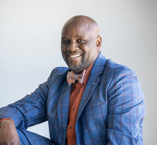 A smiling man in a blue plaid suit, orange shirt, and bow tie sitting against a plain background.