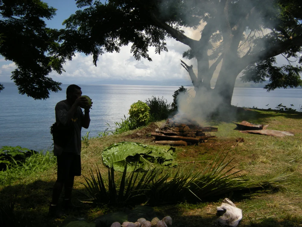 Api drinks from a coconut while tending the lovo fire.