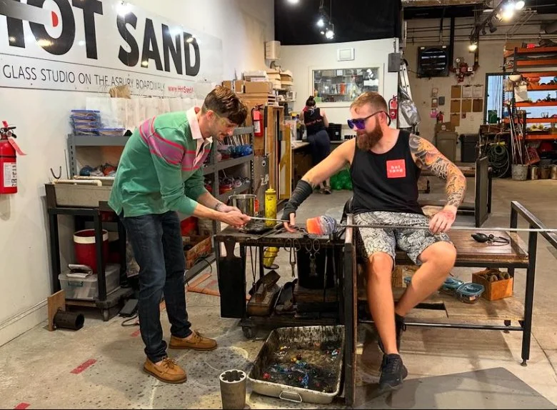 Two men working at a glass studio, one standing and shaping glass while the other sits on a bench and holds a glass rod, surrounded by tools and equipment in an industrial workshop.
