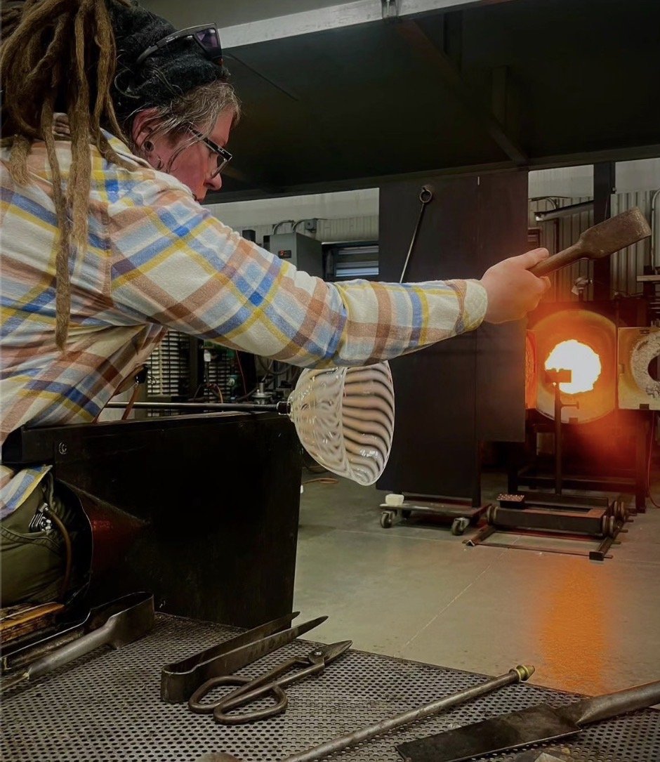 A glassblower wearing glasses and a plaid shirt is shaping molten glass with a wooden tool in a glasswork studio.