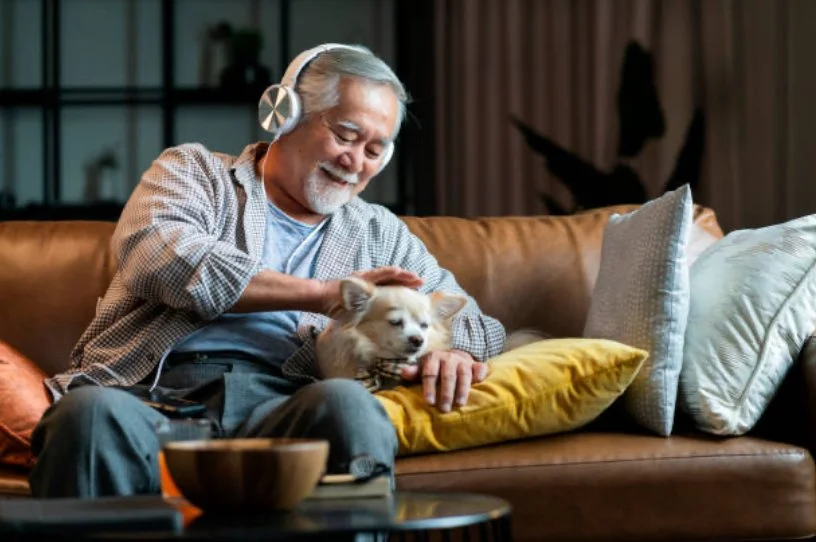 An elderly man with white hair, beard, and headphones smiling while petting a small dog on a sofa in a cozy living room.