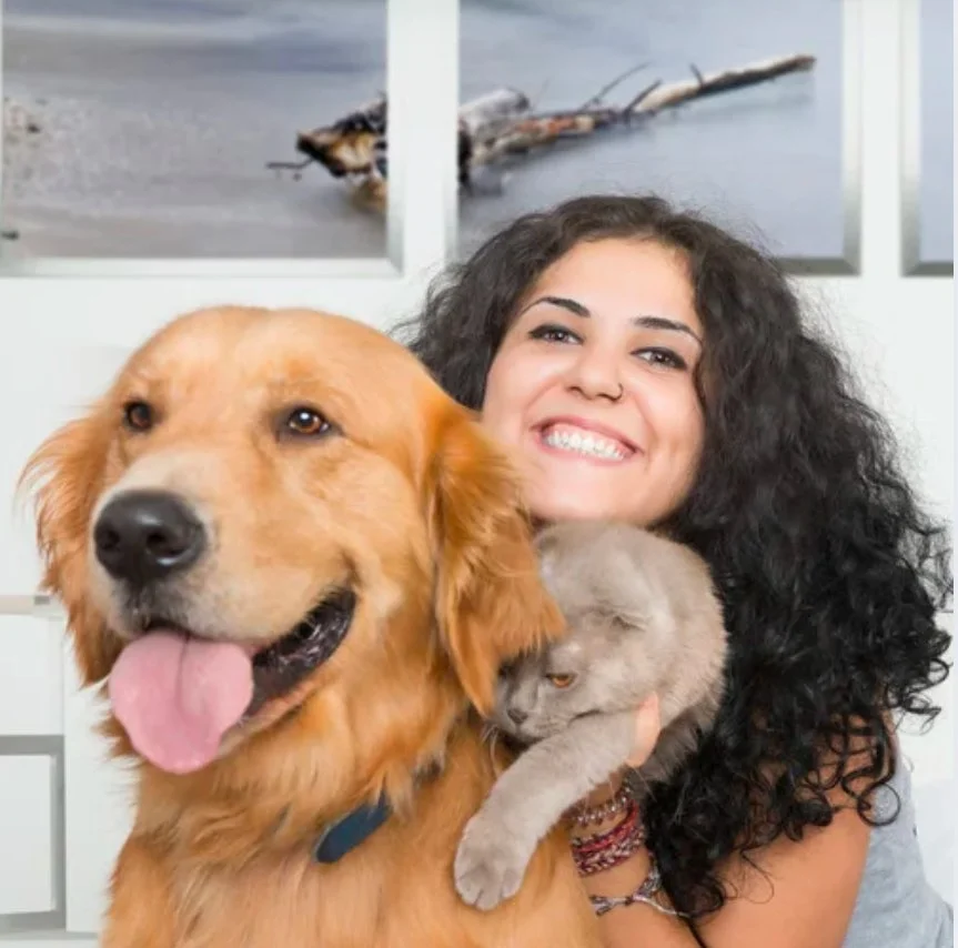 A woman with curly dark hair smiling while cuddling her golden retriever dog and a gray cat, with beach photos in the background.