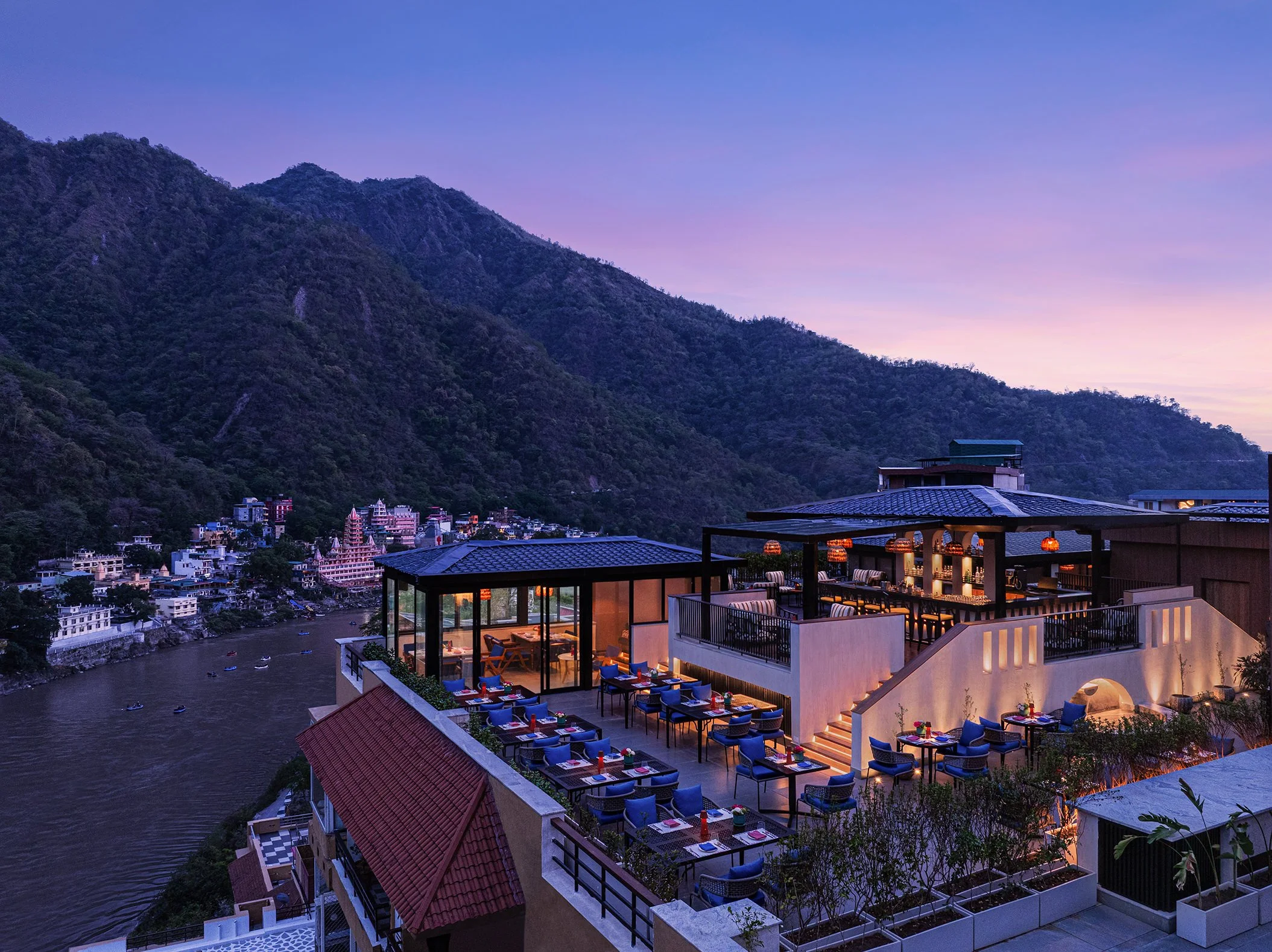 Twilight view of Aloha on the Ganges rooftop restaurant in Rishikesh, blue-cushioned dining tables overlooking the river and illuminated hillside town.