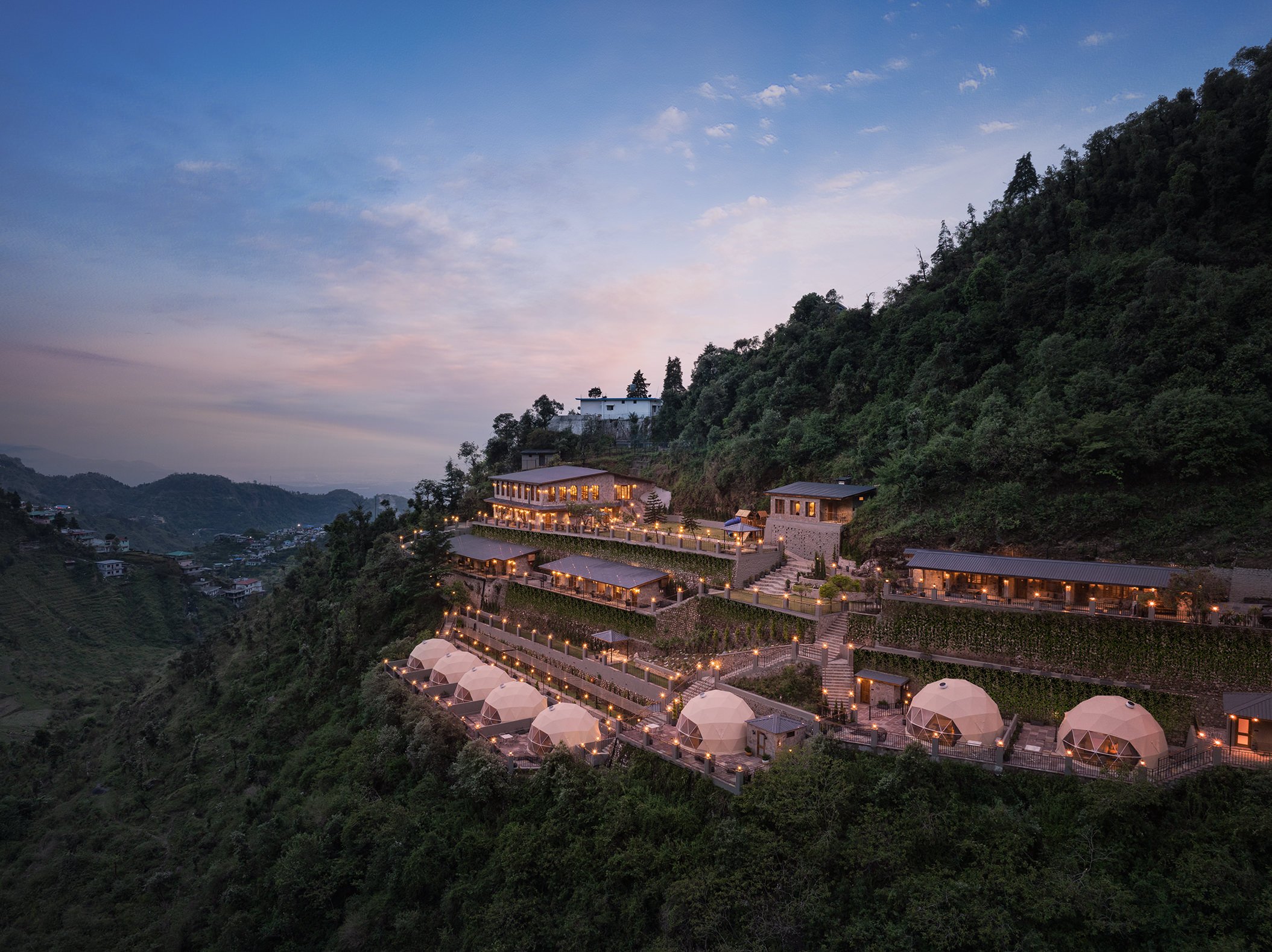 Aerial dusk view of Samsara Hill Resort by Leisure Hotels, Mussoorie, terraced dome tents and lodges lit up on a steep forested hillside overlooking the valley.