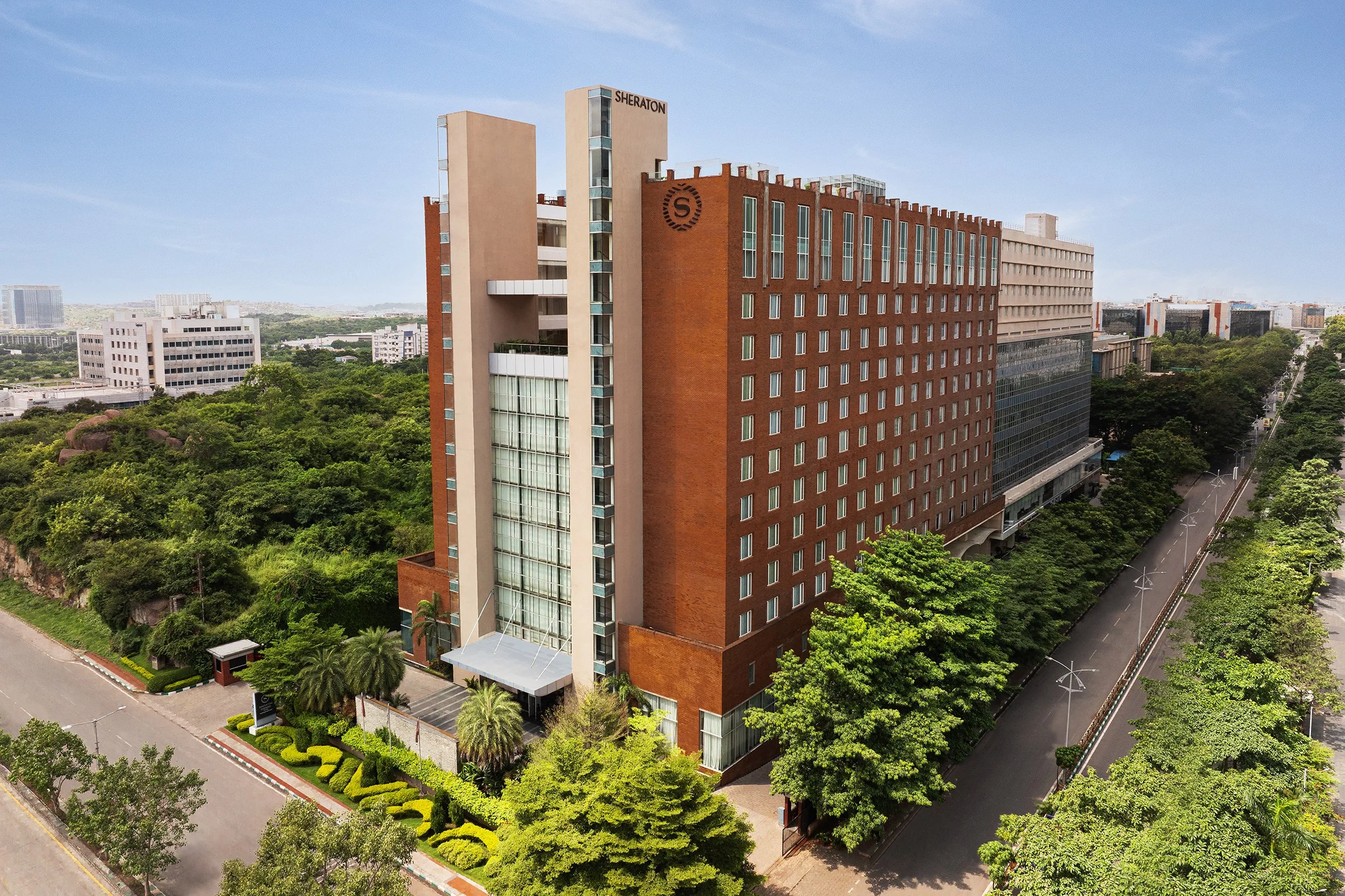 Aerial daytime view of the Sheraton Hyderabad hotel tower, red-brick façade rising above the tech corridor's greenery.