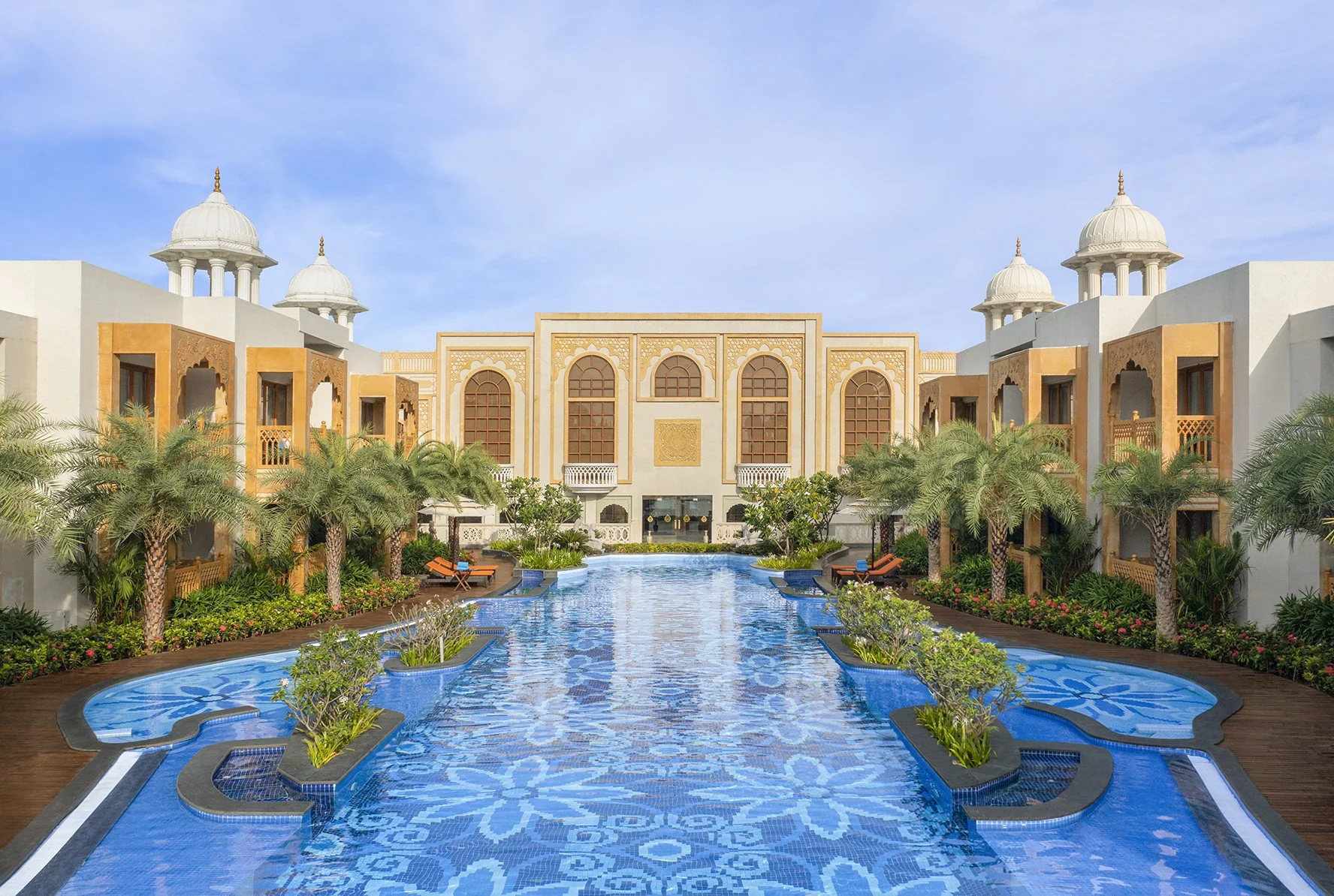 Symmetrical daytime view of Kaldan Samudra Palace's Rajasthani-style courtyard, palm-lined mosaic-tiled pool leading to an ornately arched façade with domed turrets.