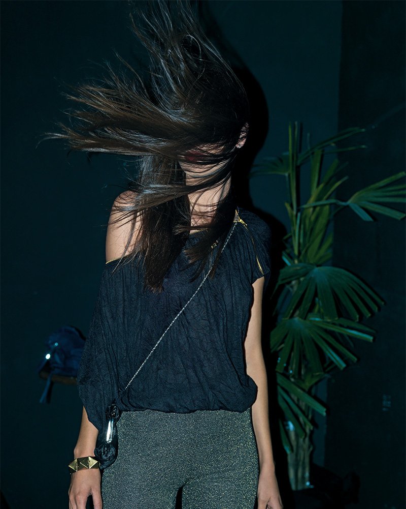 Person standing indoors with hair covering face, wearing a dark outfit with metallic pants, and a plant in the background.