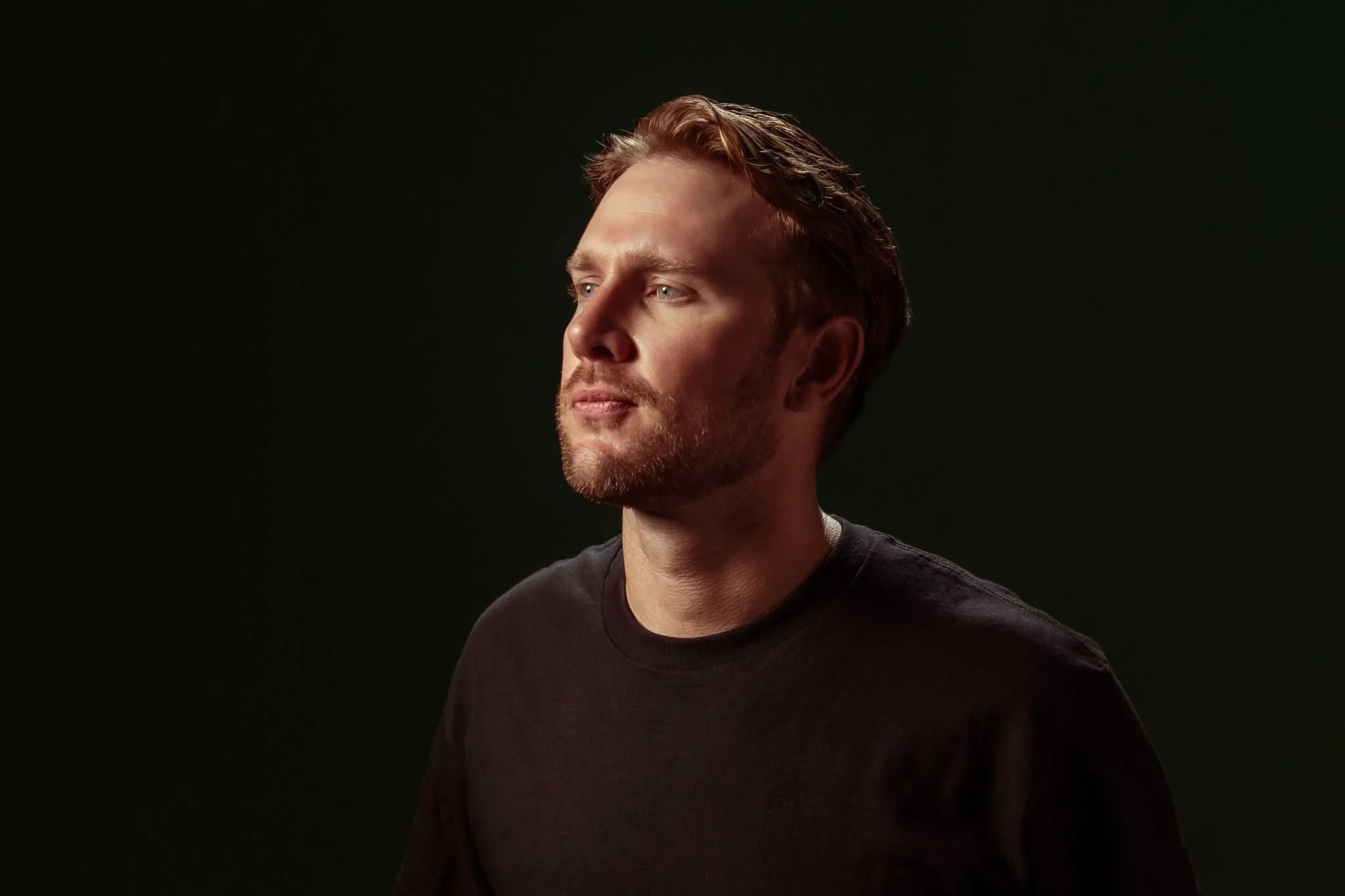 Portrait of a man with reddish-brown hair, light beard, wearing a black shirt, against a dark background.