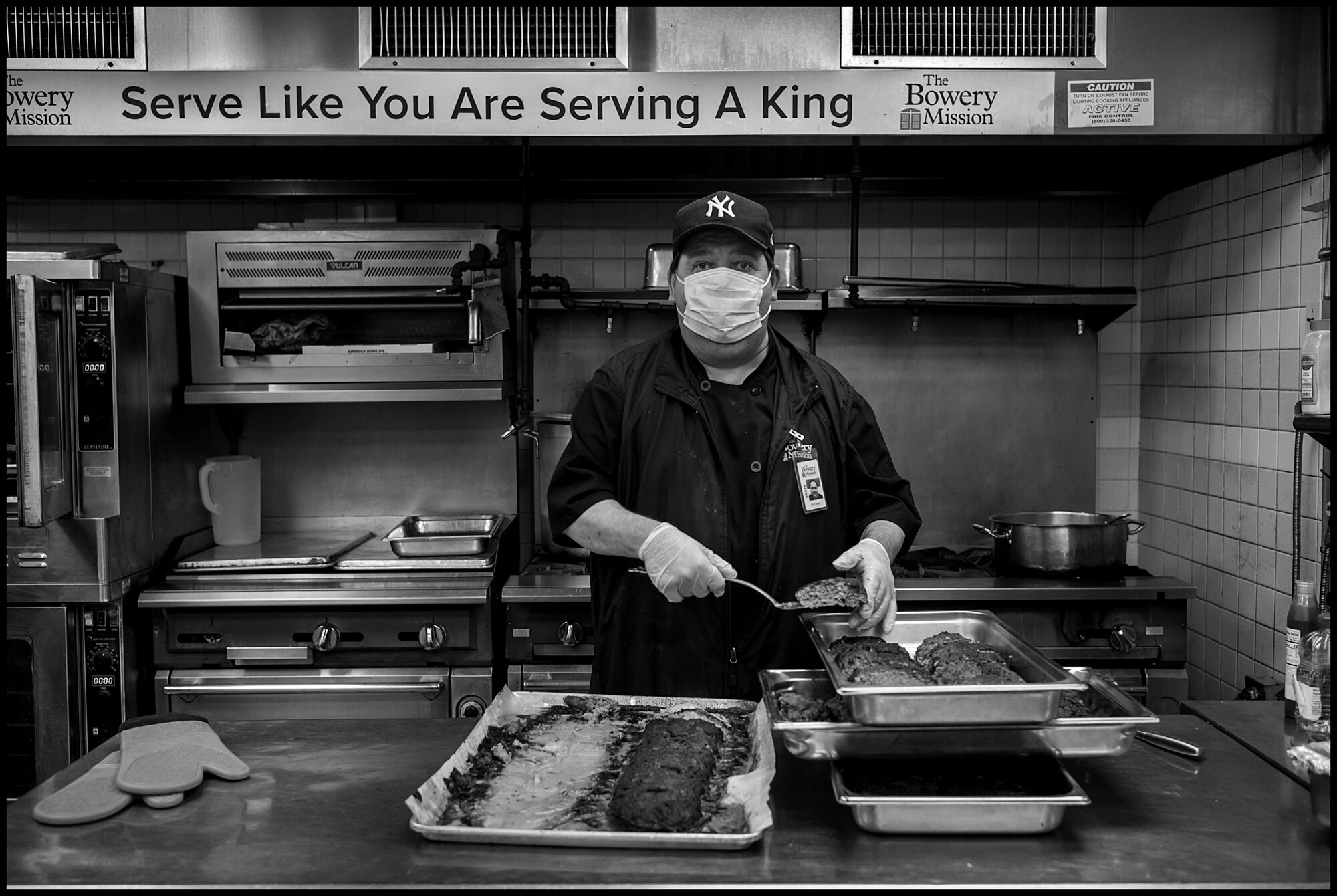  John, 58, a cook at The Bowery Mission, stands over a grill preparing food, with a sign above saying, “Serve Like You Are Serving a King”.   April 10, 2020. © Peter Turnley.   ID# 18-005 