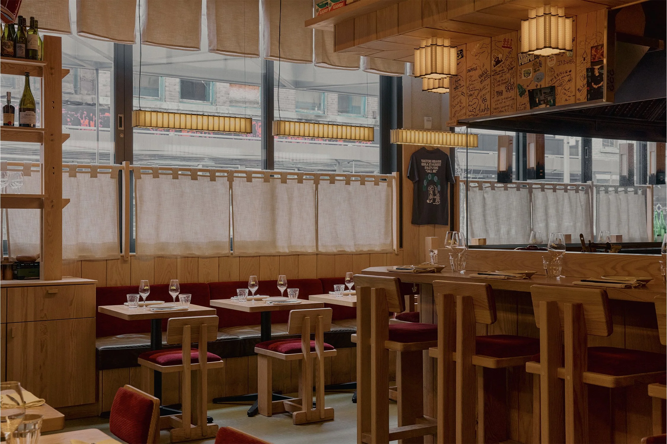 Dining area at Hibahihi with custom wood seating, linen window curtains, and suspended lantern lighting along the street facade.