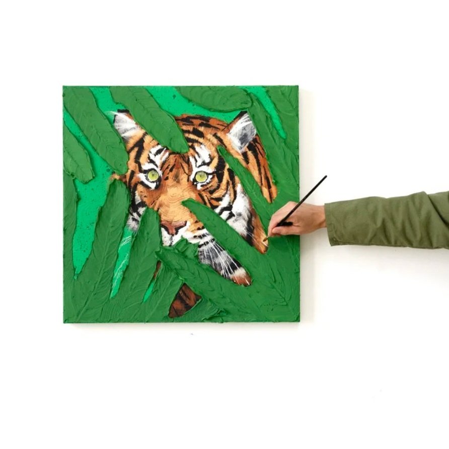 Detail image of the artist putting final touches on a painting of a tiger hidden in tropical leaves, emphasizing depth, texture, and vivid green tones by Josh Brown.