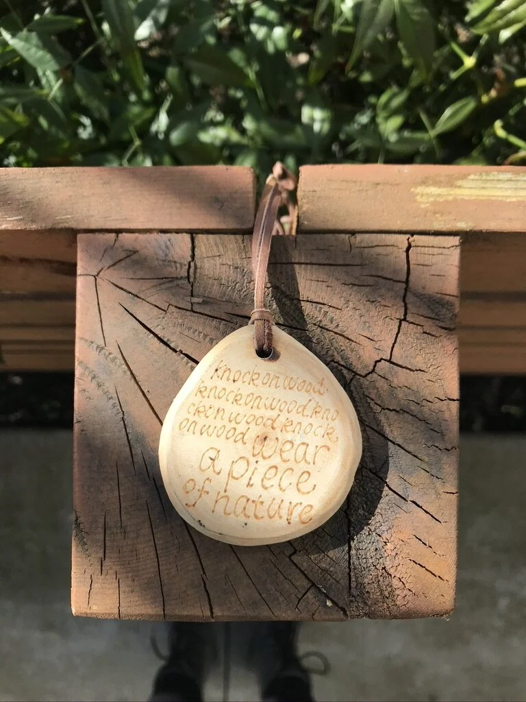 image of a wood pendant with engraved text: knock on wood, wear a piece of nature