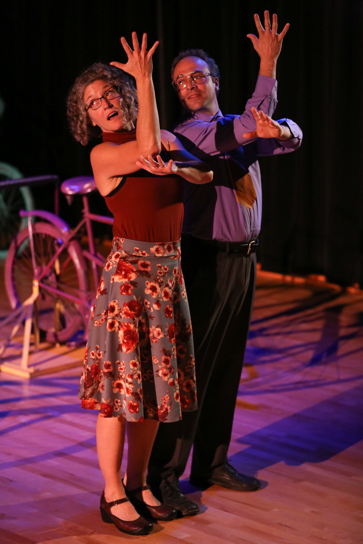 A group of teens and older adults dressed in vibrant, colorful clothing, all sitting/riding/moving around matching colorful stationary bicycles, emotions varying from curious, serious, and playful.
