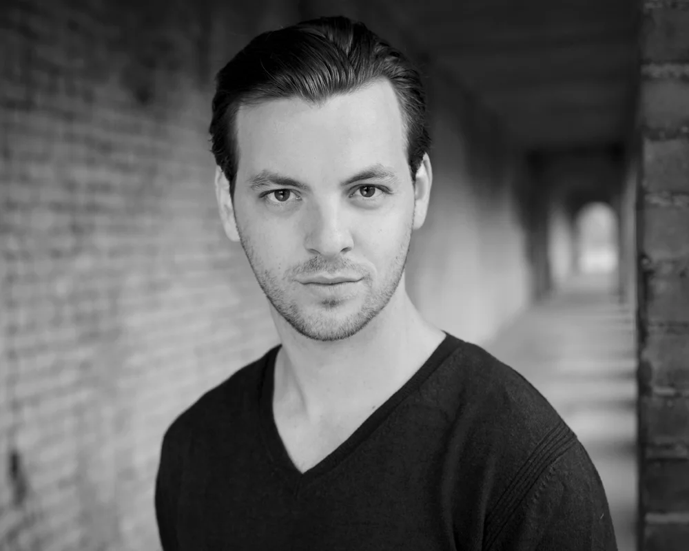 Black and white portrait of a young man with dark hair, wearing a V-neck shirt, standing outdoors under an archway with brick walls on either side.
