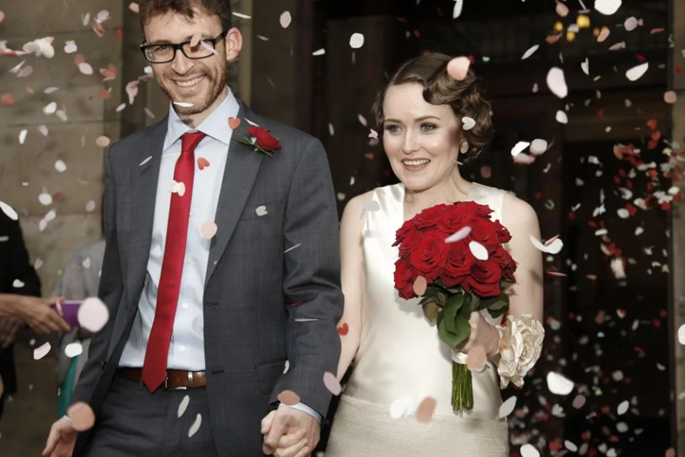 A couple at a wedding celebration, holding hands and smiling, with confetti falling around them. The groom is wearing a gray suit, white shirt, red tie, and glasses, with a red rose boutonniere. The bride is wearing a white satin dress, holding a bou